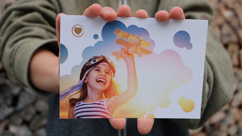Das Bild auf der Postkarte zeigt ein lachendes Mädchen beim Spiel mit einem Holzflieger, der bereit zum Abflug ist. (Bild: zVG/CHINDERNETZ AI)